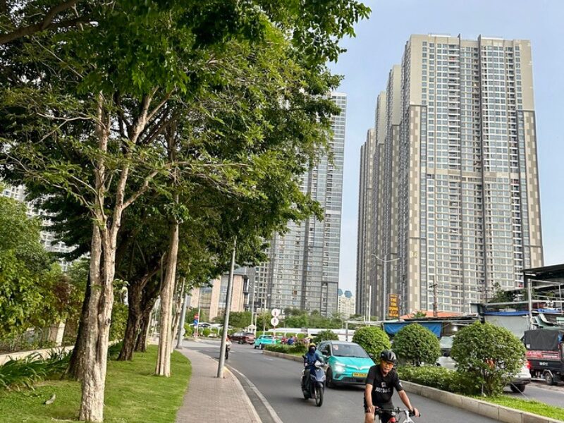 Vietnam real estate surge in HCMC: Tall apartment towers lining a bustling urban street with scooters, cyclists, and lush greenery.