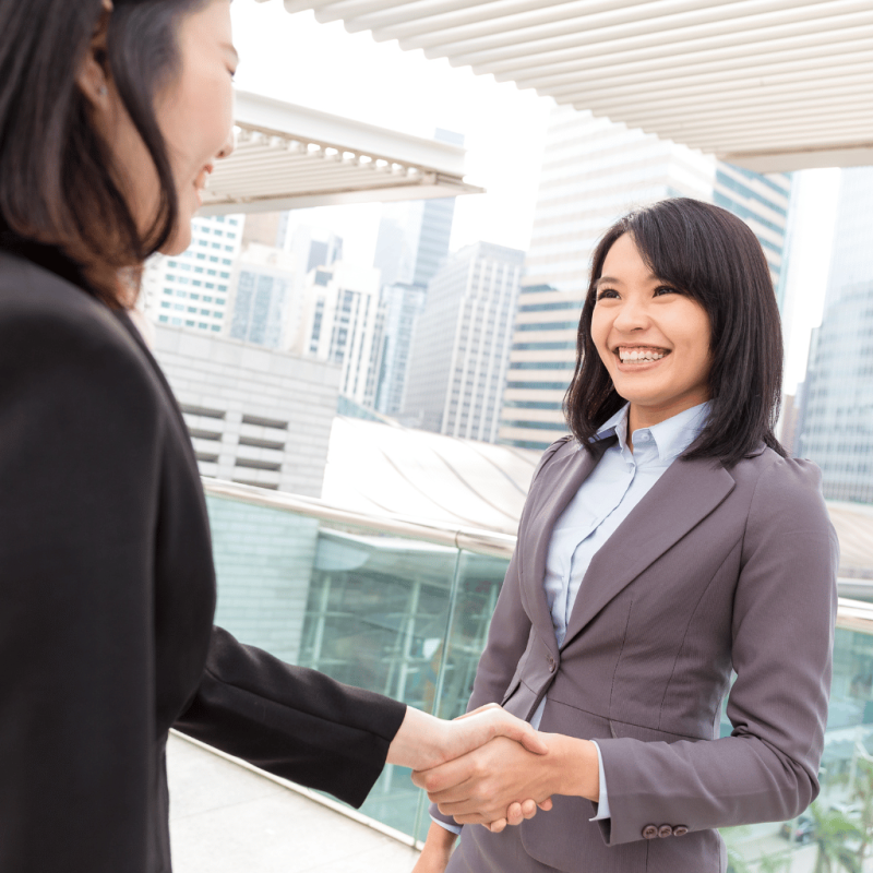 Two businesswomen shaking hands on a rooftop with a city skyline in the background, symbolizing a successful partnership in real estate investment.