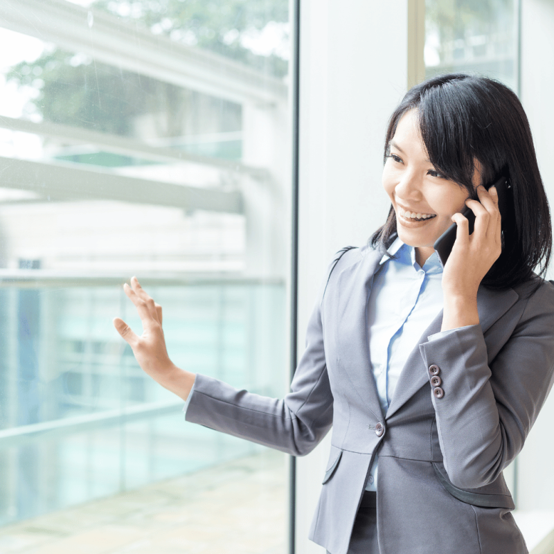 Alt Text: A professional woman in a grey suit speaking on the phone with a gesture, standing near a large office window, conducting due diligence for real estate acquisition.