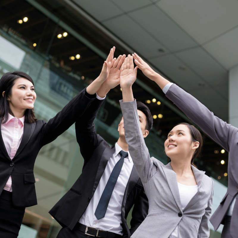 A team of professionals in business attire giving high-fives in a modern office, celebrating successful real estate development and profit maximization.