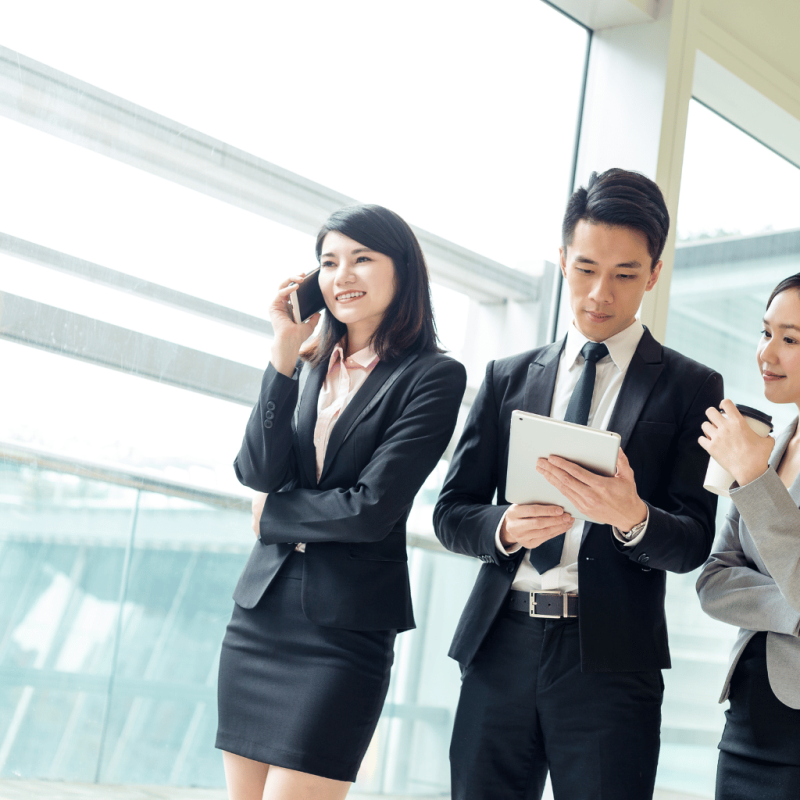 Group of professional consultants in business attire discussing real estate options, with one on a phone call, another reviewing a tablet, and a third holding a coffee cup, in a modern office setting.